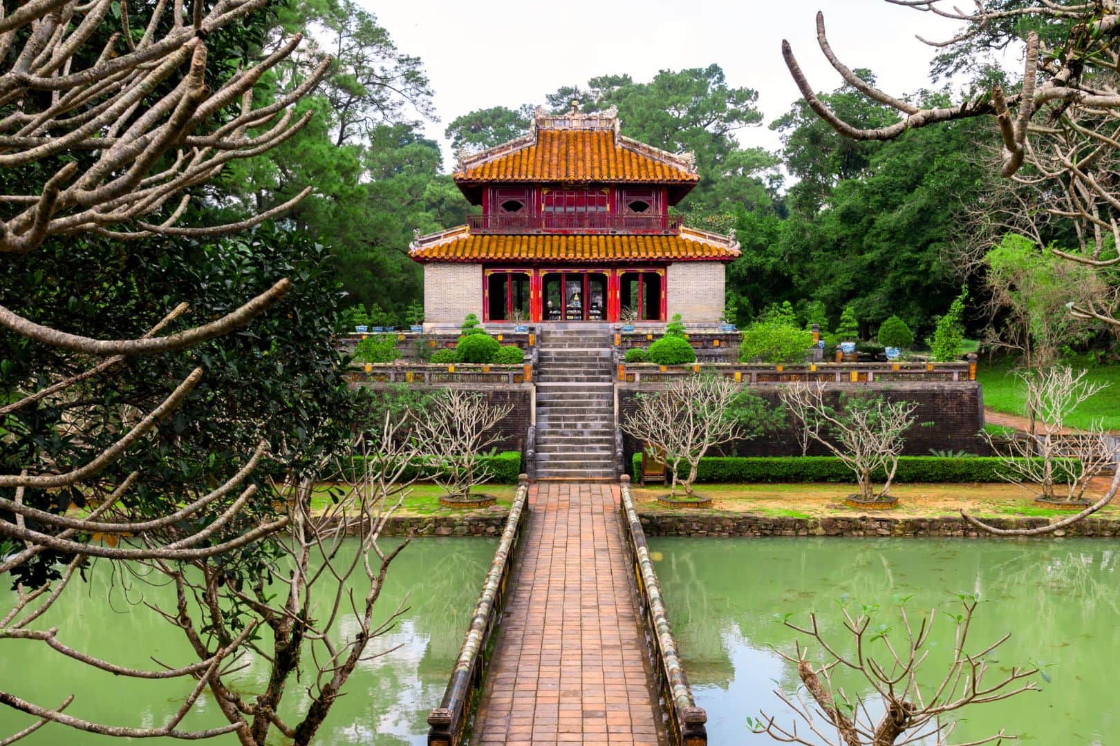 A two-storey pavilion at a royal tomb in Hue, reached by a brick bridge across a green pond